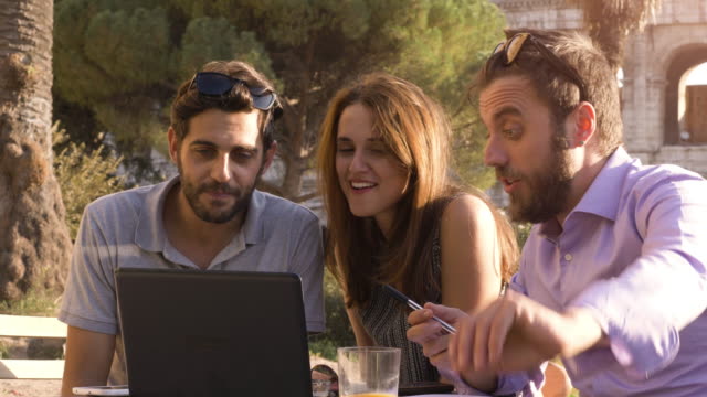 Three-young-people-working-together-on-a-project-with-laptop-and-tablet-brainstorming-writing-talking-and-researching-sitting-at-bar-restaurant-table-in-front-of-colosseum-in-rome-at-sunset