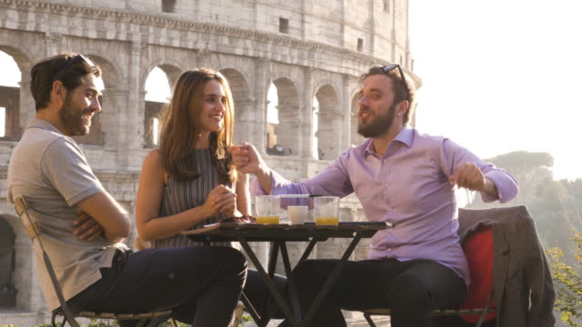 Three-young-friends-having-fun-laughing-telling-stories-and-jokes-with-exagerated-gestures-sitting-at-bar-restaurant-table-in-front-of-colosseum-in-rome-at-sunset