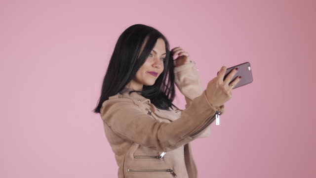 Lächelnde-glücklich-Brünette-Frau-in-rosa-Jacke-Selfie-auf-Smartphone-über-farbigen-Hintergrund-machen.-4k