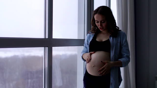 Una-feliz-mujer-embarazada-se-encuentra-en-un-gran-ventanal-situado-a-una-altitud-alta.-Él-toca-su-estómago-con-las-manos-y-sonriendo-comunica-con-el-bebé-en-el-vientre