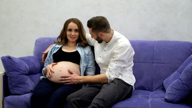 A-married-couple-is-sitting-at-home-on-the-couch-waiting-for-the-birth-of-a-child.-The-man-stroked-her-hand-across-her-belly-and-kissing-the-belly-of-his-pregnant-wife