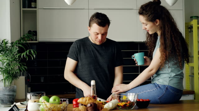 Glückliches-junges-Paar-Kochen-und-plaudern,-während-glücklich-Mann-Schneiden-von-Gemüse-zum-Frühstück-in-der-Küche-zu-Hause.-Beziehung-und-Familie-Konzept
