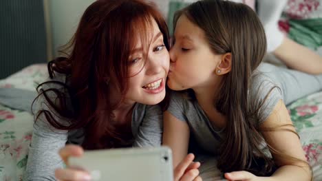 Glückliche-Mutter-und-Mädchen-nehmen-Selfie-Foto-mit-Smartphone-Kamera-und-viel-Spaß-beim-Grimassieren-beim-Sitzen-im-kuscheligen-Bett-zu-Hause
