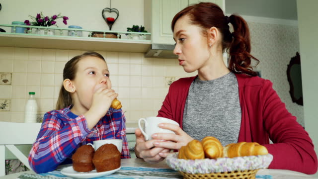 Cheerful-mother-and-cute-daughter-having-breakfast-eating-muffins-and-croissants-talking-at-home-in-modern-kitchen.-Family,-food,-home-and-people-concept