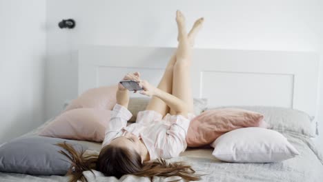 carefree-young-brunette-is-laying-on-bed-,-rising-legs-on-headboard-and-typing-messages-and-posting-in-internet-using-mobile-phone