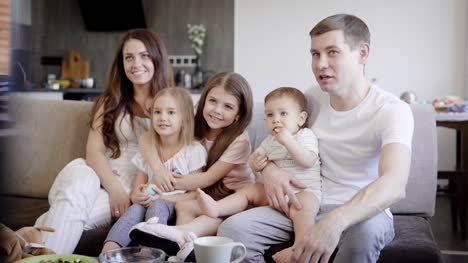 glückliche-Familie-Paar-mit-drei-kleinen-Töchtern-auf-einer-Couch-im-Wohnzimmer-sitzen-und-Fernsehen-tagsüber