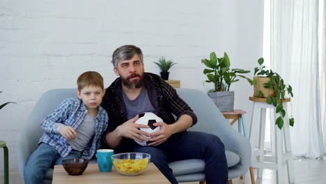 Konzentrierte-Vater-und-Sohn-beobachten-Fußballspiel-im-Fernsehen-zu-Hause,-jubeln,-feiern-Sieg-mit-fünf-und-Snacks-zu-essen.-Glückliche-Familie-und-Sport-Konzept.