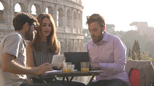 Three-people-working-together-on-a-project-with-laptop-and-tablet-writing-talking-and-researching-sitting-at-bar-restaurant-table-in-front-of-colosseum-in-rome-at-sunset
