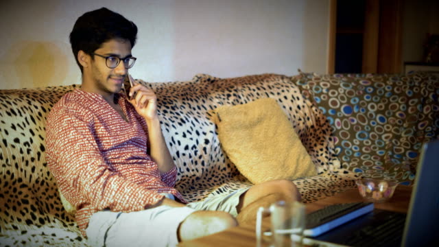 Indian-Man-In-Glasses-Discussing-By-Phone-Business-Questions-Sitting-On-A-Sofa-In-Front-Of-A-Laptop