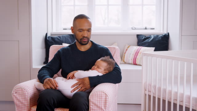 Stressed-New-Father-Sitting-In-Chair-Holding-Sleeping-Baby-Girl-In-Nursery-At-Home