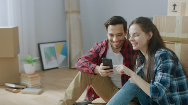 Feliz-pareja-joven-sentada-en-el-piso-del-apartamento-recién-alquilado-/-adquirido-uso-Smartphone.-Cajas-de-cartón-desempaquetados-y-muebles-cubiertos-en-el-moderno-brillante-dulce-hogar.