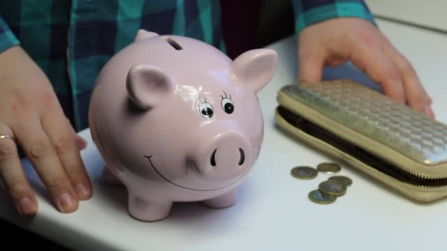 On-the-table-there-is-a-piggy-bank-in-the-form-of-a-pink-pig.-Lying-wallet-and-scattered-coins.-A-man-collects-coins-from-the-table-and-puts-them-in-a-piggy-bank.
