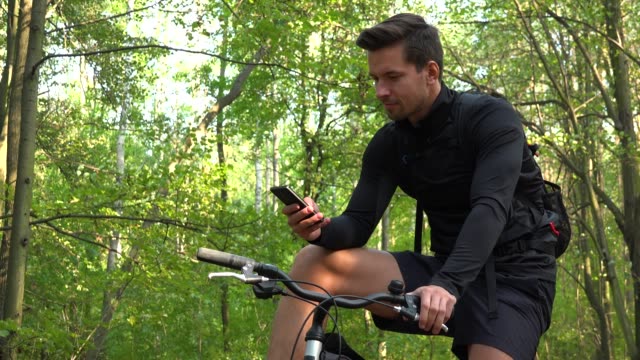 Un-ciclista-joven-y-guapo-se-sienta-en-su-bicicleta-en-un-bosque-y-trabaja-en-una-vista-de-teléfono-inteligente-desde-abajo