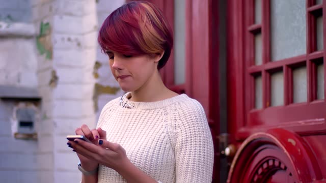 Portrait-of-young-caucasian-pink-haired-girl-joyfully-working-with-smartphone-on-red-door-background.