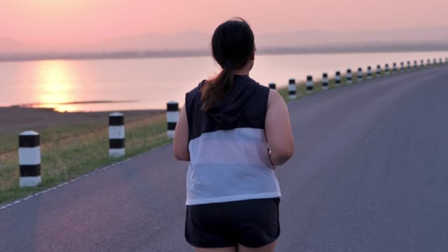 Übergewichtige-asiatische-Frauen-joggen-am-frühen-Morgen-im-Sonnenlicht-auf-der-Straße.-Konzept-des-Gewichtsverlustes-mit-Bewegung-für-die-Gesundheit.-Langsame-Bewegung,-Rückansicht