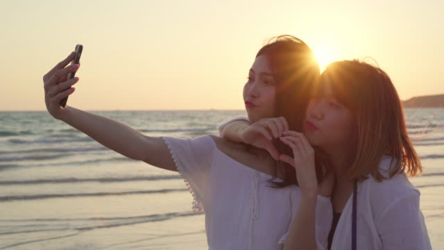 Young-Asian-lesbian-couple-using-smartphone-taking-selfie-near-beach.-Beautiful-women-lgbt-couple-happy-relax-enjoy-love-moment-when-sunset-in-evening.-Lifestyle-lesbian-couple-travel-on-beach-concept