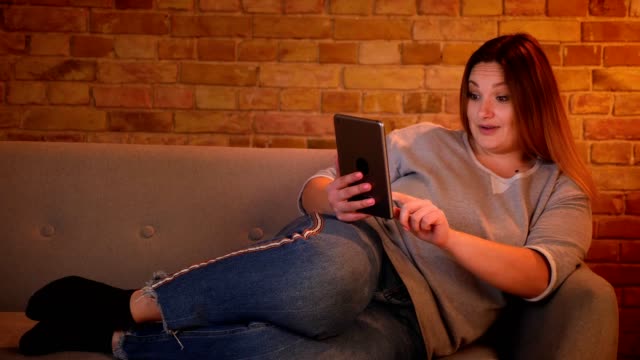 Portrait-of-relaxed-plus-size-long-haired-model-lie-on-sofa-chatting-joyfully-on-tablet-in-cozy-home-atmosphere.
