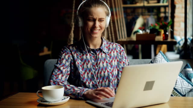 Fröhliche-junge-Frau-in-Kopfhörern-hören-Musik-mit-Laptop-im-Café