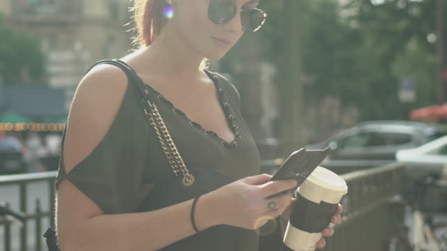Young-attractive-woman-writing-a-text-message-on-her-smartphone-at-the-subway-exit-in-street,-holding-her-coffee-in-other-hand,-during-sunny-summer-afternoon-in-Paris.-Trendy-and-cool,-tilt-up.