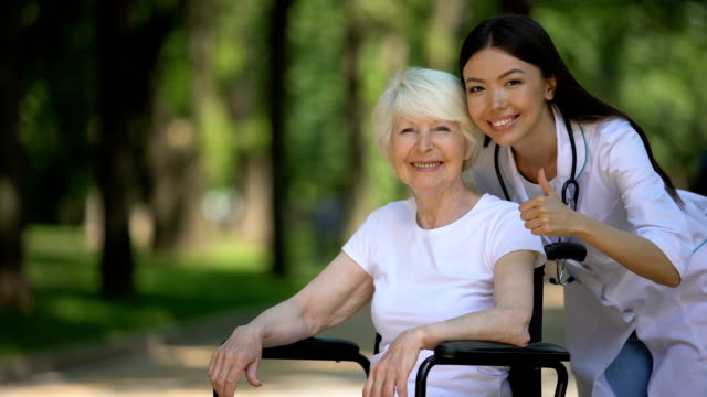 Nurse-and-elderly-woman-in-wheelchair-smiling-at-camera-and-showing-thumbs-up