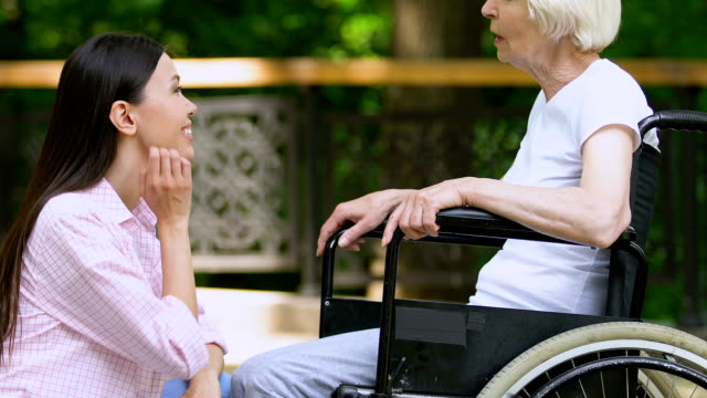 Lächelnde-weibliche-Freiwillige-im-Gespräch-mit-älteren-Frau-im-Rollstuhl,-Unterstützung