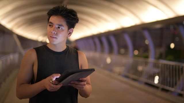Porträt-von-jungen-stattlichen-asiatischen-Mann-denken,-während-mit-digitalen-Tablet-im-Freien-in-der-Nacht