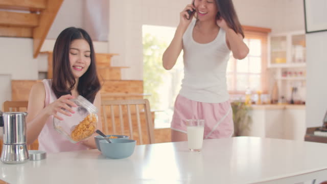 Pareja-lesbiana-asiática-usando-teléfono-móvil-hablar-con-un-amigo-mientras-desayuna-en-la-cocina-en-la-mañana.
