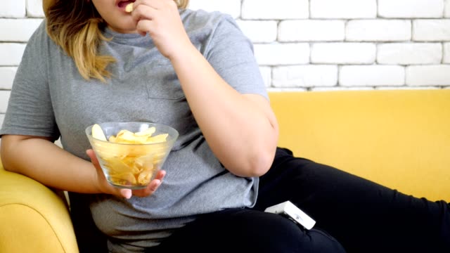 Mujer-gorda-comiendo-papas-fritas-en-el-sofá-y-viendo-la-televisión-en-casa