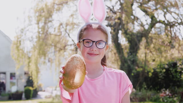 Portrait-of-girl-wearing-bunny-ears-on-Easter-egg-hunt-outdoors-at-home-holding-up-chocolate-egg-to-camera-and-smiling---shot-in-slow-motion