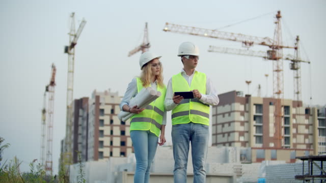 Two-colleagues-of-engineers-a-man-and-a-woman-discussing-a-drawing-and-a-tablet-computer-on-the-background-of-buildings-under-construction-and-cranes,-a-woman-talking-on-the-phone-with-the-boss.