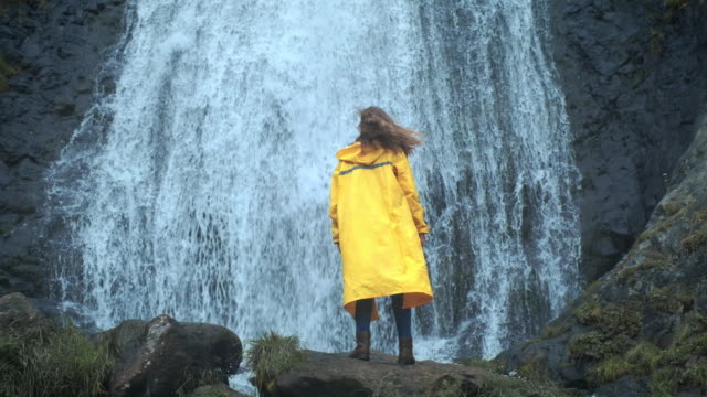 Junge-Mädchen-Wanderer-in-einem-gelben-Regenmantel-geht-zu-einem-Wasserfall-im-Hochland,-hebt-die-Hände-nach-oben,-genießt-Die-Natur-und-das-Leben.-Reisen-in-den-Bergen,-Abenteuer-in-Reise.-Lifestyle-Konzept