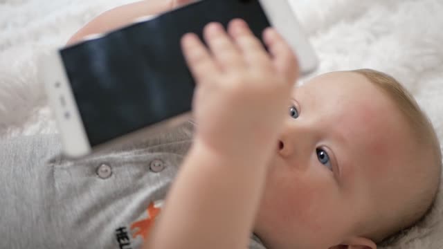 Cute-baby-holds-phone-in-hands.-The-child-plays-with-cell-phone,-examines-it,-tugs-in-his-hands-and-tries-to-eat-smartphone-putting-it-in-his-mouth