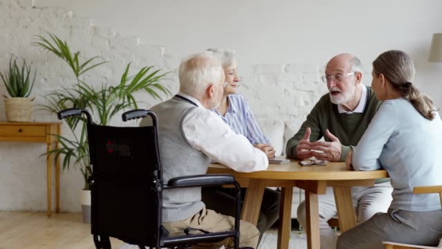 Gruppe-von-vier-älteren-Rentnern,-zwei-Männern-und-zwei-Frauen,-die-am-Tisch-sitzen-und-im-Gemeinschaftsraum-des-Pflegeheims-sprechen.-Senior-Mann-in-Brille-erzählen-Geschichte-zu-Freunden,-Tracking-Schuss