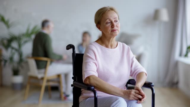 Tracking-shot-of-unhappy-disabled-senior-woman-in-wheelchair-thinking-and-looking-at-camera-with-sadness-in-nursing-home,-other-aged-patients-in-background