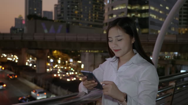Vista-lateral-de-la-joven-mujer-asiática-sonriente-usando-teléfonos-inteligentes-en-el-centro-de-la-ciudad-moderna-en-la-noche-de-Bangkok-Tailandia.-Comunicación-Concept-Technology-por-teléfono-móvil