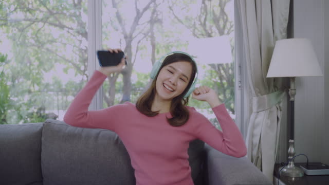 Junge-asiatische-Frau-singt-mit-glücklich,-während-mit-Handy-und-Kopfhörer-im-Wohnzimmer.
