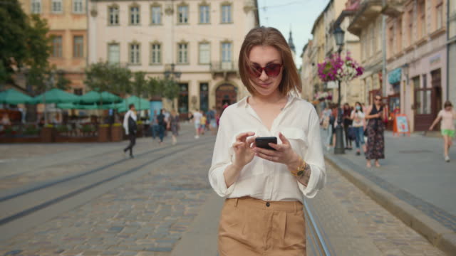 A-smiling-girl-is-walking-and-texting-on-her-smartphone.-She's-wearing-sunglasses.-People-are-walking-in-the-background.-4K