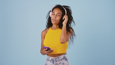 Afro-american-girl-is-choosing-a-song-on-her-mobile-phone-and-listening-it-through-headphones.-Smiling-and-dancing,-posing-on-blue-background