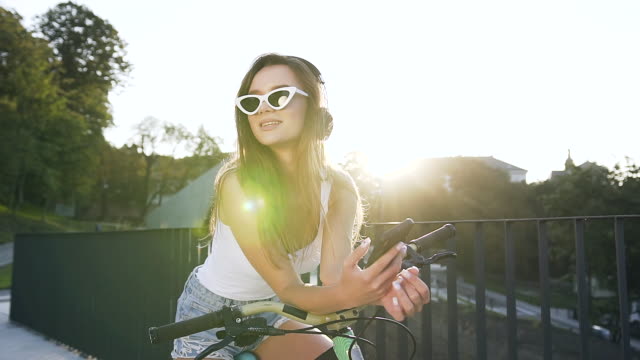 Junge-kaukasische-Frau-in-trendigen-Sonnenbrille-mit-langen-Haaren-sitzt-auf-dem-Fahrrad-und-hört-Musik-in-Kopfhörern-durch-die-Smartphone-App-in-den-Lichtern-der-Sonne-in-der-Stadt