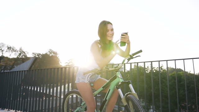 Portrait-of-attractive-smiling-girl-in-with-long-hair-sits-on-bicycle-and-something-watching-in-smartphone-app-in-summer-day-at-city