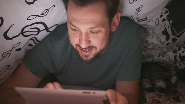 Close-up-of-smiling-face-of-a-young-bearded-and-mustashed-man,-using-digital-tablet-under-blanket,-in-his-bed,-at-night.-His-yorkshire-terrier-is-lying-next-to-him
