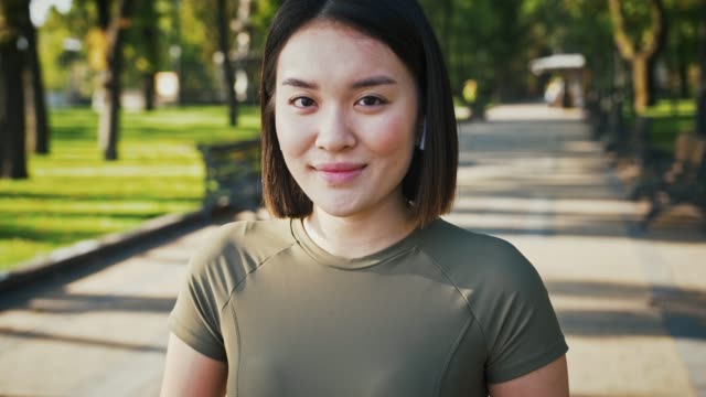 Outdoor-portrait-of-young-asian-woman,-listening-to-music-in-airpods-and-smiling-to-camera-in-park,-tracking-shot