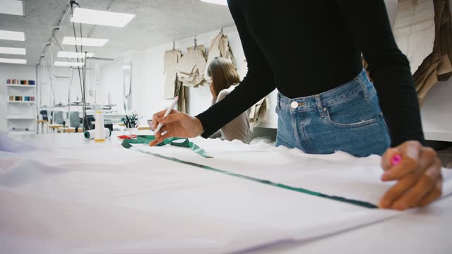 Sewing-workshop,-unknown-woman-taking-measurements-of-fabric-by-tape-measure,-colleague-clarifying-order-details-by-mobile-phone