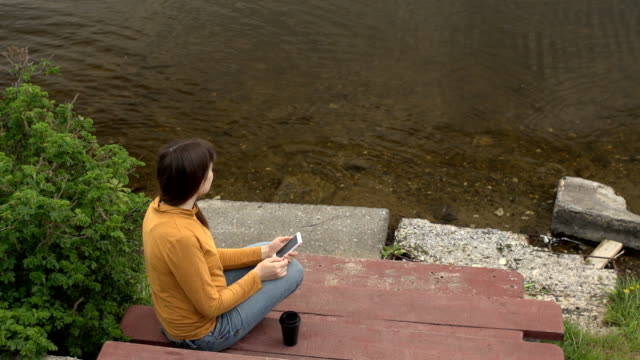 Eine-junge-Frau-genießt-eine-Telefon-sitzen-auf-einem-Pier-am-Teich-im-Sommer.