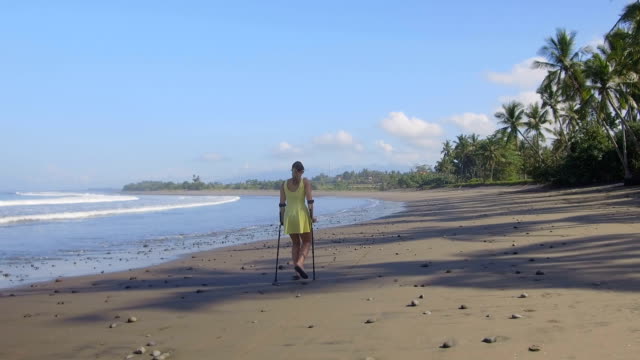 CLOSE-UP:-Woman-with-crutches-walking-down-the-sandy-beach-on-tropical-island