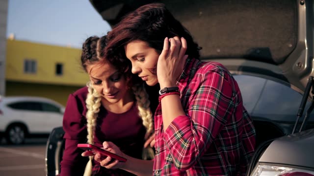 Closeup-view-of-two-young-attractive-girlfriends-looking-at-the-smartphone,-checking-pictures-while-sitting-inside-of-the-open-car-trunk-in-the-parking-by-the-shopping-mall-during-sunny-day