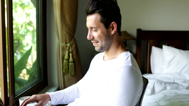 Young-Man-Using-Laptop-Computer-Happy-Smiling-Guy-Chatting-Online-Over-Big-Window-With-Tropical-Garden-View