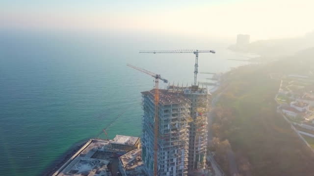 Aerial-city-view.-Construction-of-a-high-rise-skyscraper-on-the-ocean-by-two-cranes.-Flight-counter-clockwise.-The-rays-of-the-setting-sun-fall-into-the-lens