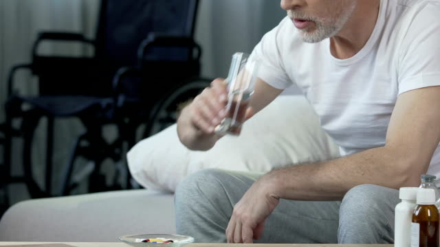 Ältere-männliche-Einnahme-Pille,-Blick-auf-Rollstuhl-stehen-im-Hintergrund,-Trauma