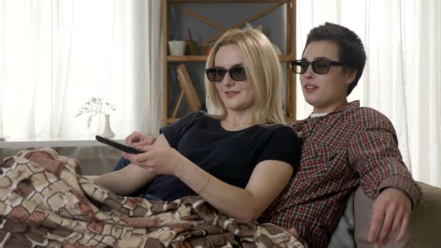 Zwei-junge-lesbische-Mädchen-sitzen-auf-der-Couch,-in-eine-warme-Decke,-bedeckt-den-3d-Film,-Fernbedienung,-lachen.-60fps